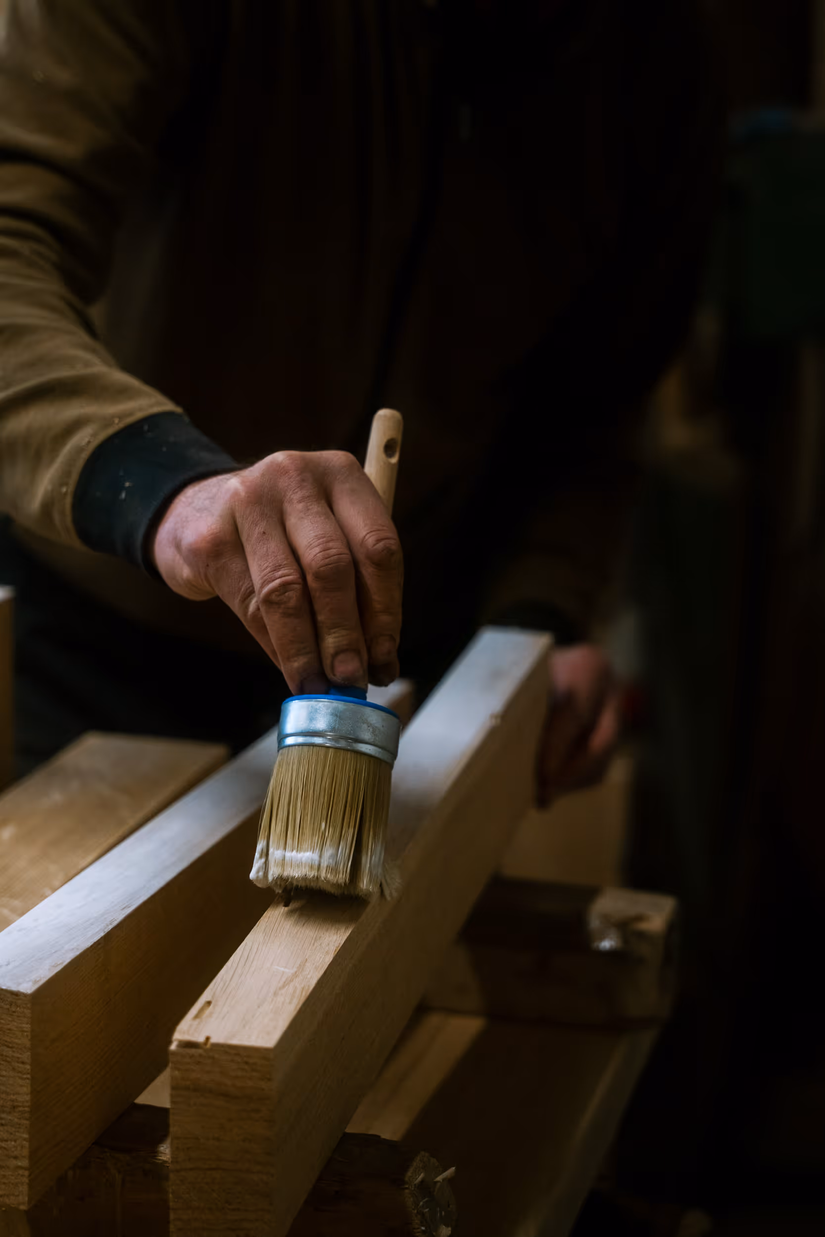 Ręka trzymająca pędzel nakładająca farbę na drewnianą deskę w warsztacie stolarskim.