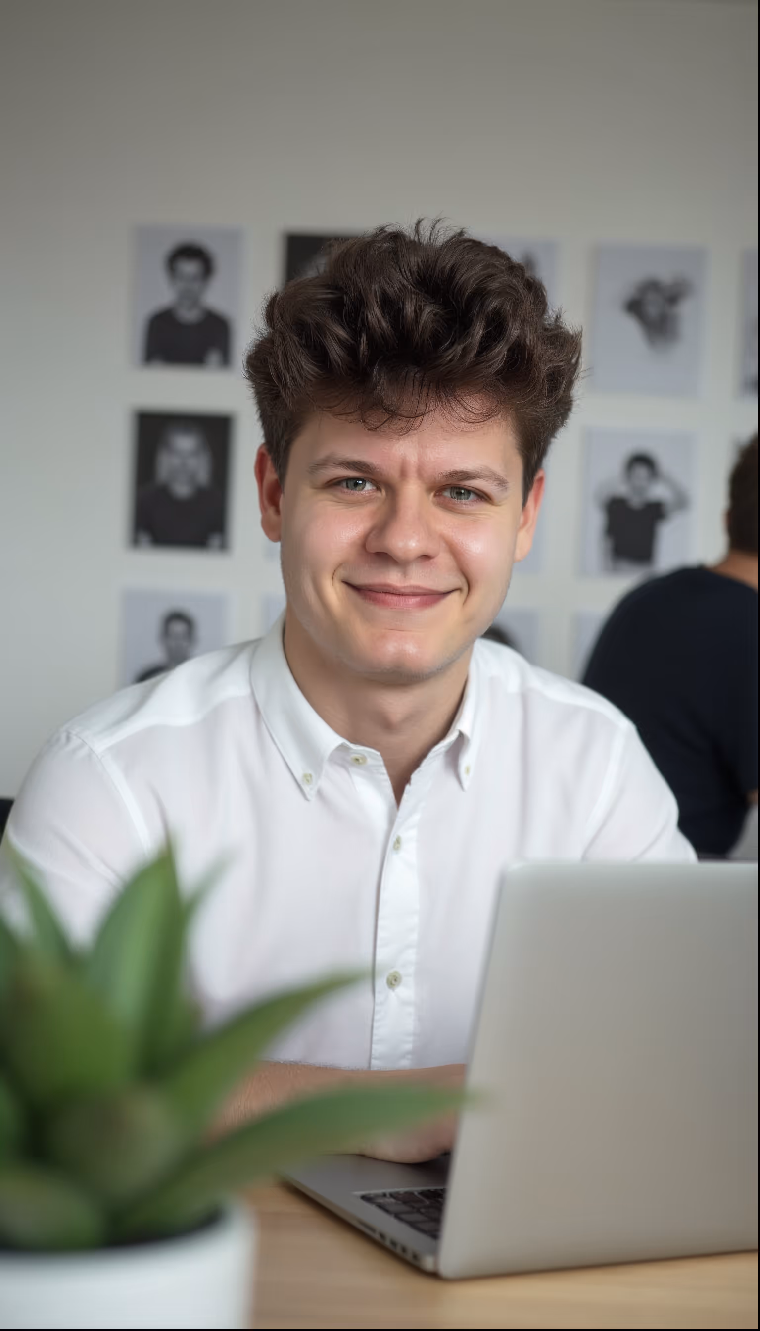 Junger Mann im weißen Hemd sitzt mit Laptop im Büro und lächelt in die Kamera, an der Wand hängen Schwarz-Weiß-Porträts.