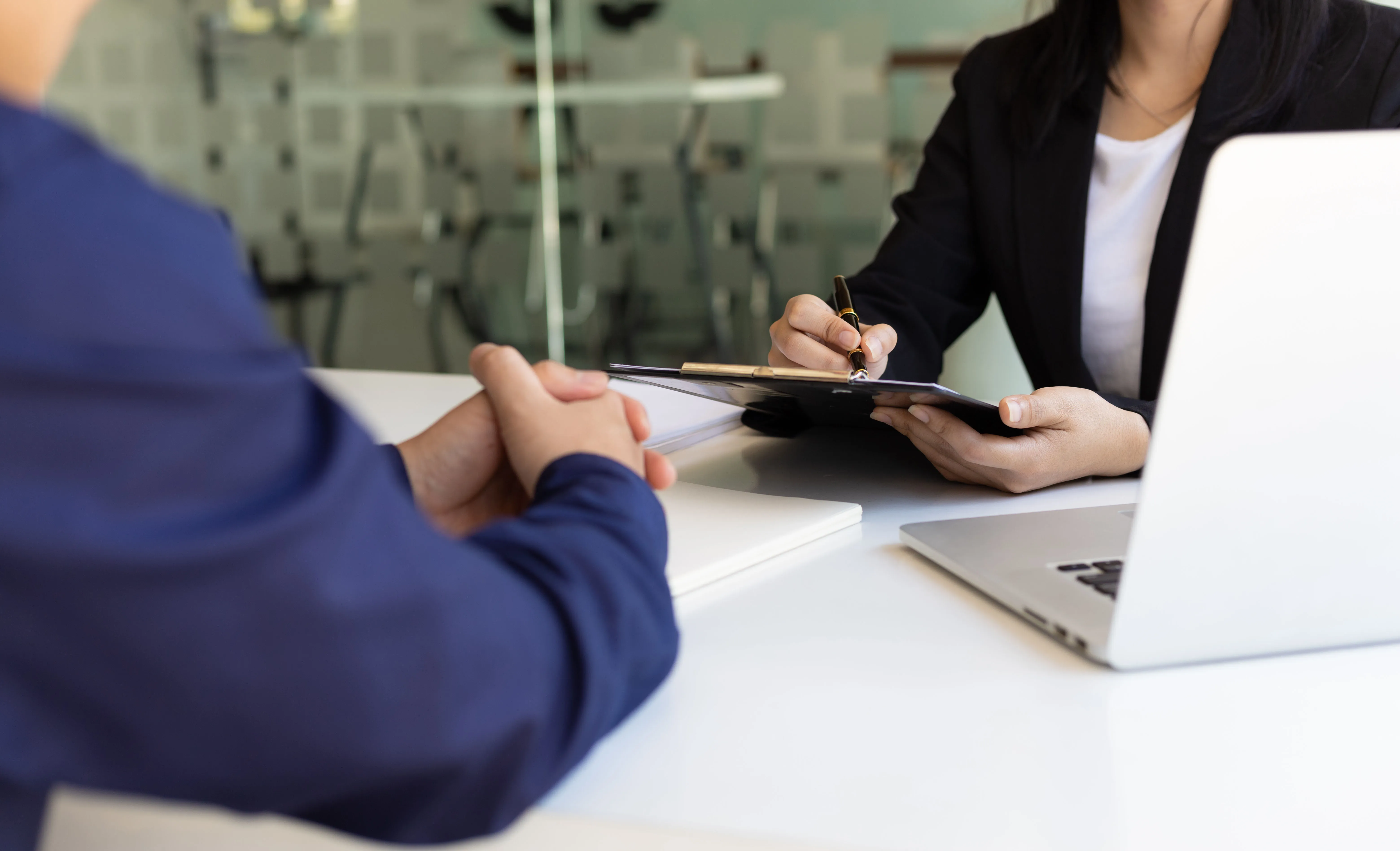 Beraterin schreibt auf einem Klemmbrett während eines Gesprächs mit einem Kunden über einen Tisch mit Laptop.