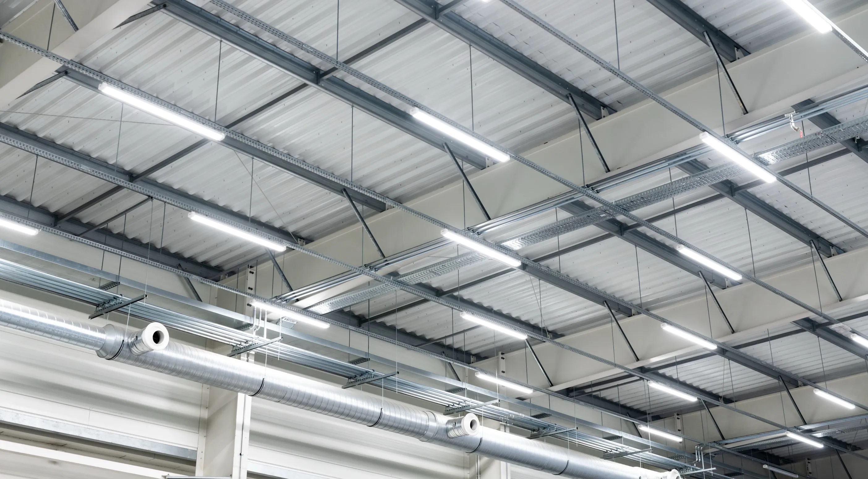 Beleuchtete Decke einer Lagerhalle mit hängenden LED-Leuchten und Lüftungsschläuchen aus Metall.
