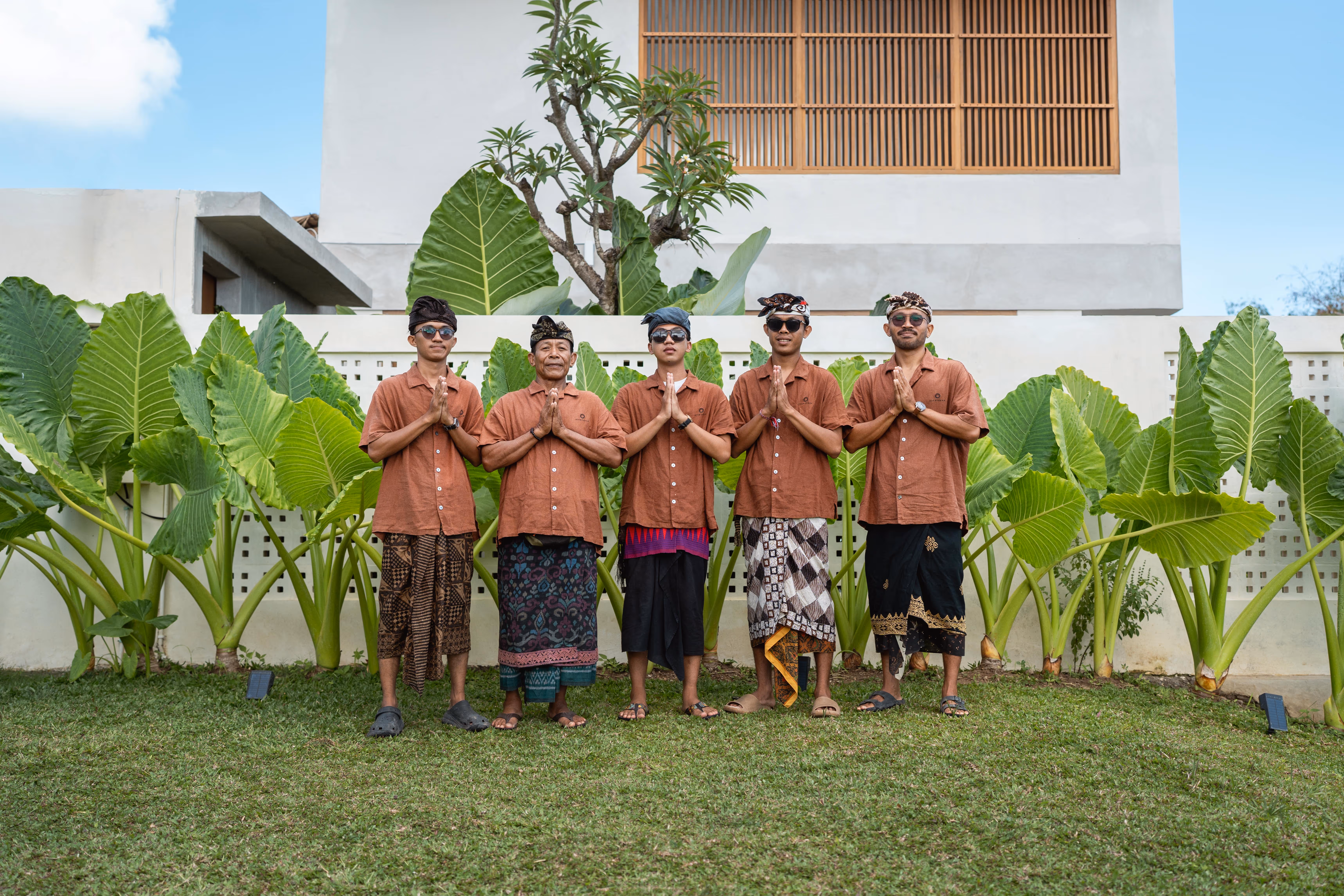Team Mizu Village. Five men in traditional Balinese attire standing on grass with hands in a prayer gesture in front of large green leaves and a white wall.