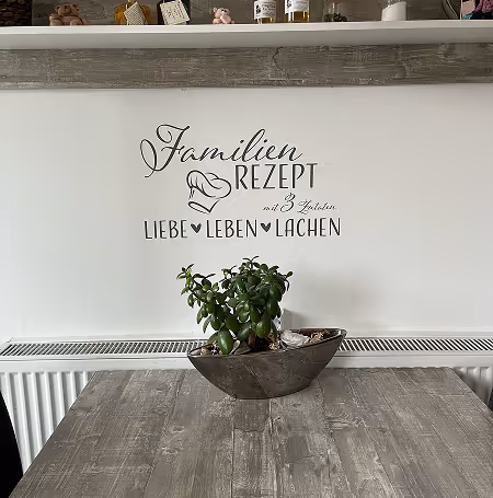 Wand mit Aufschrift 'Familienrezept mit 3 Zutaten Liebe, Leben, Lachen' über einem blauen Sitzkissen und Holztisch mit grünem Pflanzendekor.