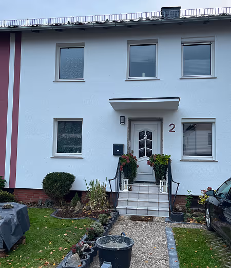 Eingang zu einem weißen Reihenhaus mit Hausnummer 2, Treppe mit Geländer und Blumen, Auto rechts geparkt.