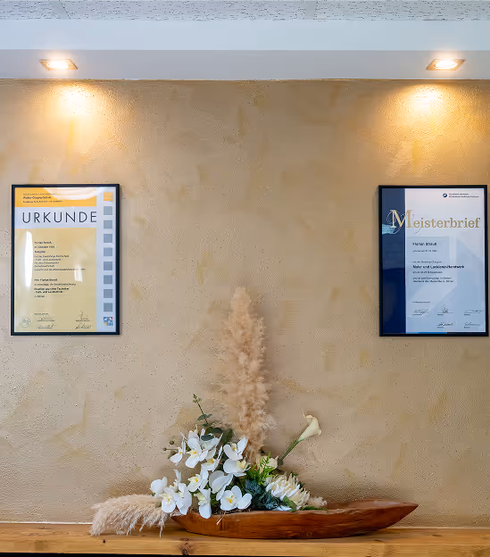 Zwei gerahmte Zertifikate hängen an einer beigen Wand über einem Holztisch mit einem dekorativen Holztablett, das weiße Blumen und Trockenpflanzen enthält.