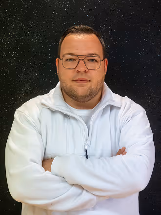 Porträt eines Mannes mit Brille und weißem Pullover vor schwarzem, leicht glitzerndem Hintergrund.