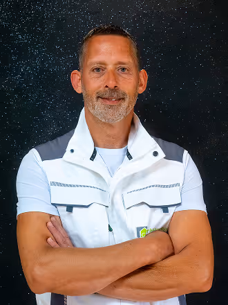 Mittelaltiger Mann mit kurzen Haaren und grau meliertem Bart, verschränkte Arme tragend, vor schwarzem, sternenähnlichem Hintergrund.