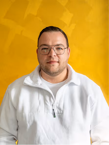 Porträt eines Mannes mit Brille und weißem Pullover vor gelbem Hintergrund.