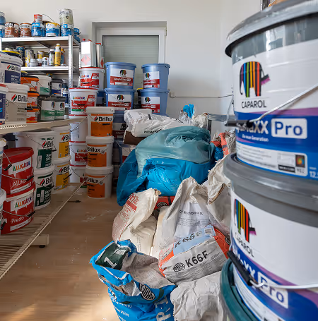Raum mit mehreren Farbdosen und Säcken mit Baumaterialien, darunter Caparol-Farbdosen und Brillux-Produkte.