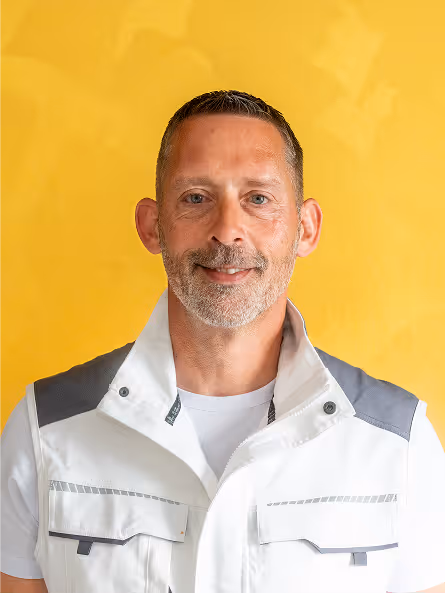 Porträt eines lächelnden Mannes mit grauem Bart und kurzem Haar, der eine weiße Arbeitsjacke vor gelbem Hintergrund trägt.