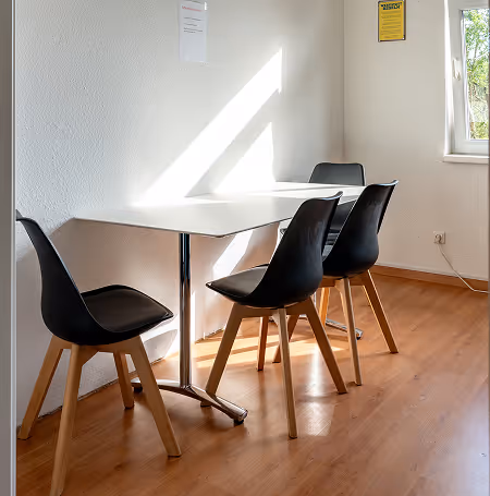 Dreiergruppe schwarzer Stühle mit Holzbeinen an einem weißen Tisch in einem hellen Raum mit Holzfußboden und Fenster.