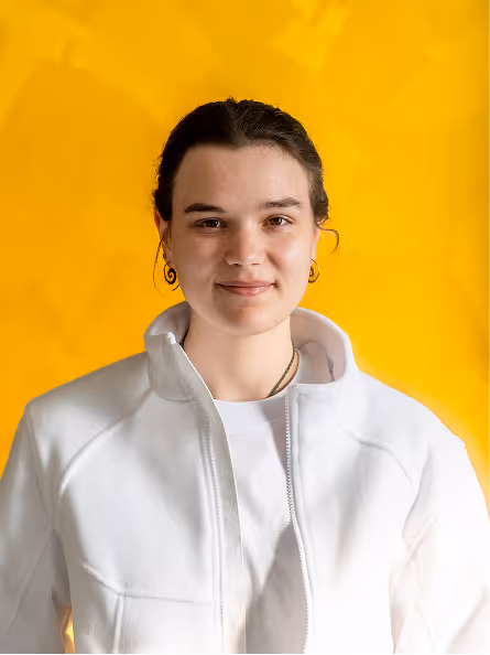 Junge Frau mit dunklem Haar, die einen weißen Reißverschluss-Pullover vor einem gelben Hintergrund trägt und lächelt.