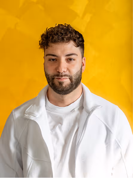 Junger Mann mit dunklem lockigem Haar und Bart trägt weißen Pullover vor gelbem Hintergrund.