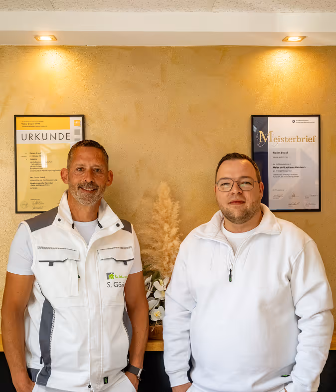 Zwei Männer in weißer Kleidung stehen vor einer weißen Wand mit zwei gerahmten Zertifikaten.