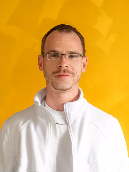 Mann mit Brille, kurzem Haar und weißem Pullover vor gelbem Hintergrund.