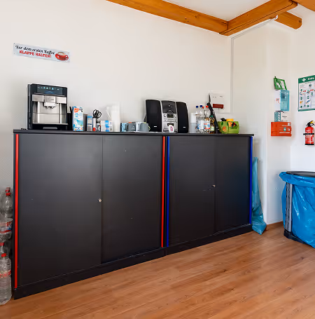 Küche mit schwarzem Schrank, Kaffeevollautomat, Radio, Wasserflaschen und Feuerlöscher an weißer Wand.