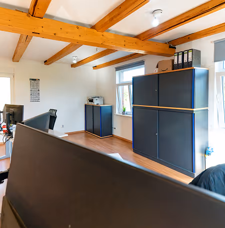 Helles Büro mit sichtbaren Holzbalken an der Decke, schwarzen Aktenschränken, Schreibtischen und Computern.