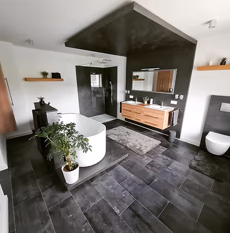 Modern bathroom with a freestanding white bathtub, dark tiled floor, wall-mounted double wooden vanity, large mirror, and potted plant.