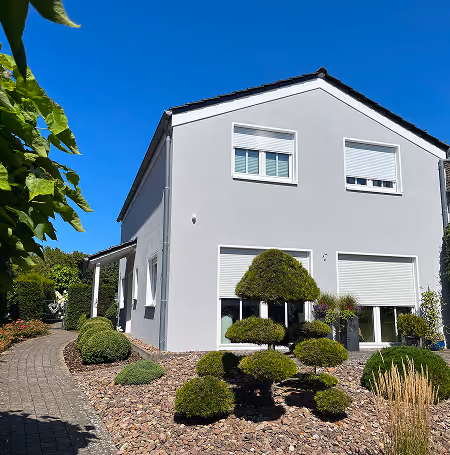 Modern weißes Haus mit Rollläden und gepflegtem Garten mit geformten Büschen unter klarem blauem Himmel.