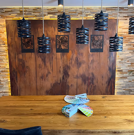 Moderner Holztisch mit Farbfächern darauf vor einer Wand mit Holzvertäfelung und sechs schwarzen hängenden Lampen.