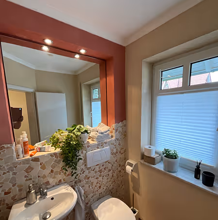 Kleines Badezimmer mit Waschbecken, Toilette, Fenster mit Jalousie, Pflanzen und Badeutensilien.