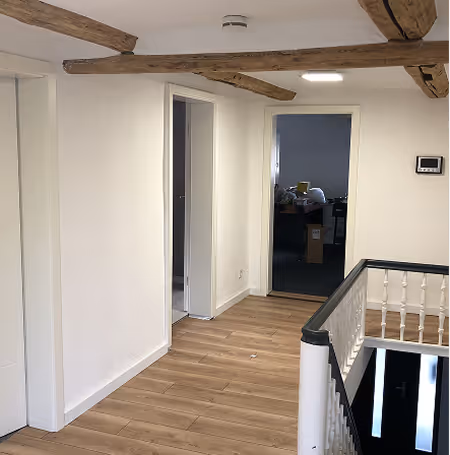 Flur mit Holzfußboden, weißen Wänden, sichtbaren Holzbalken an der Decke und offenem Durchgang zu einem dunkleren Raum mit Schreibtisch.