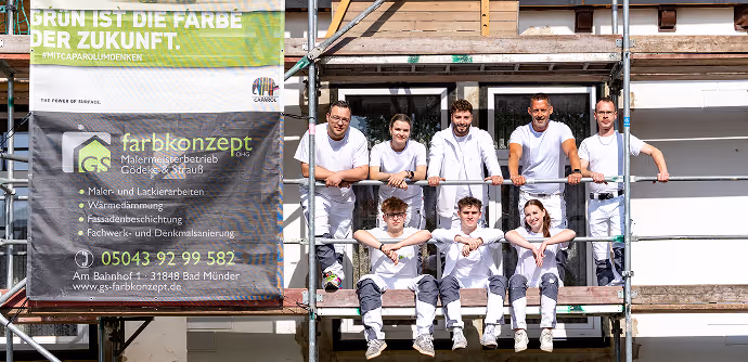 Sieben Maler in Arbeitskleidung sitzen und lehnen an einem Gerüst vor einem Haus unter einem Banner mit Firmeninformationen.