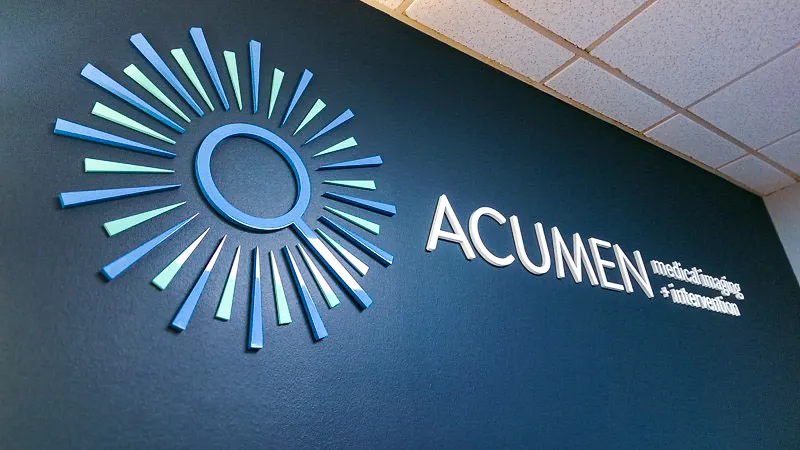Indoor Acumen sign with sunburst logo on dark wall near drop ceiling