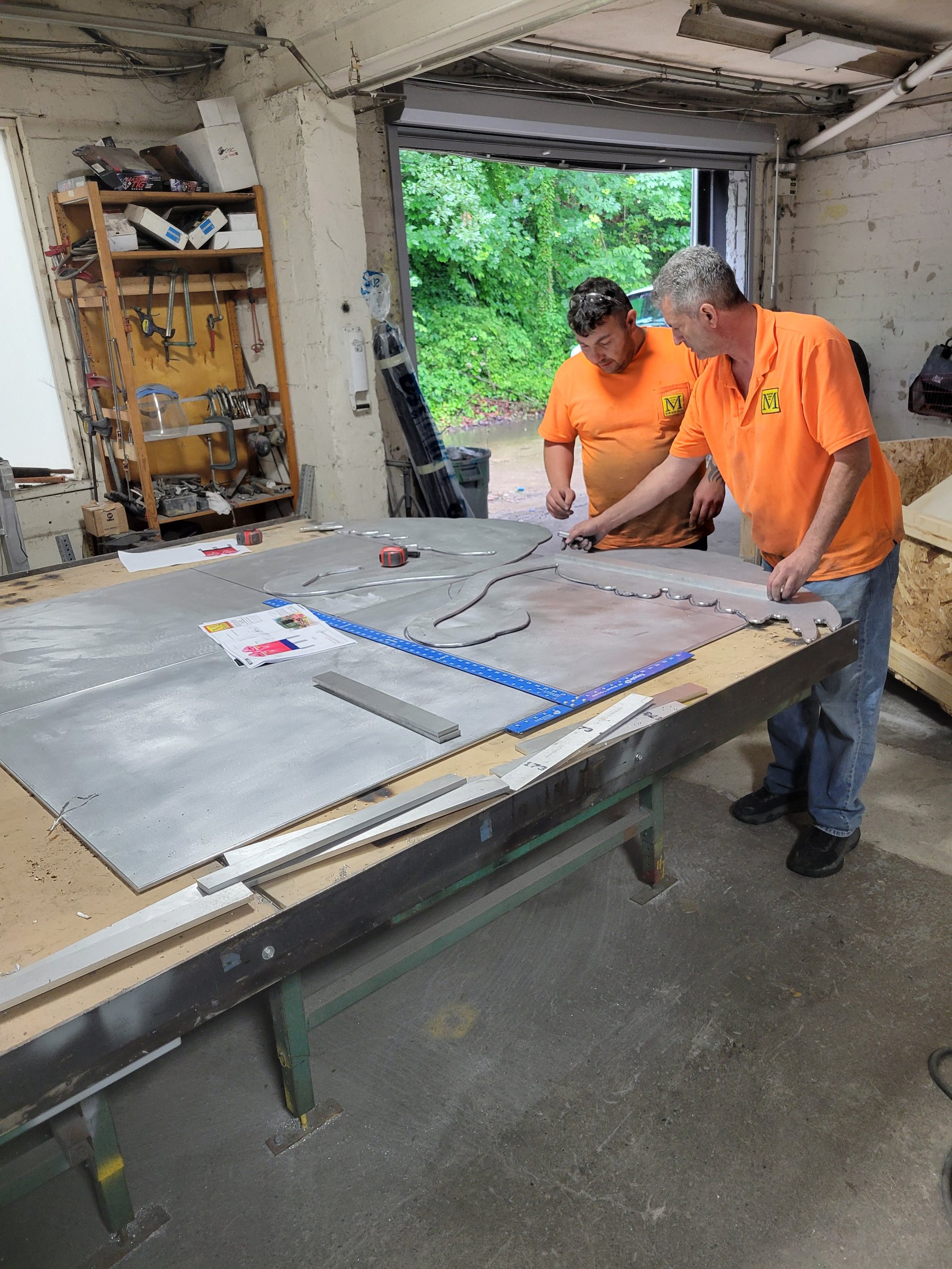 Two Martino fabricators in orange working aluminum panels at the bench with the shop doors open