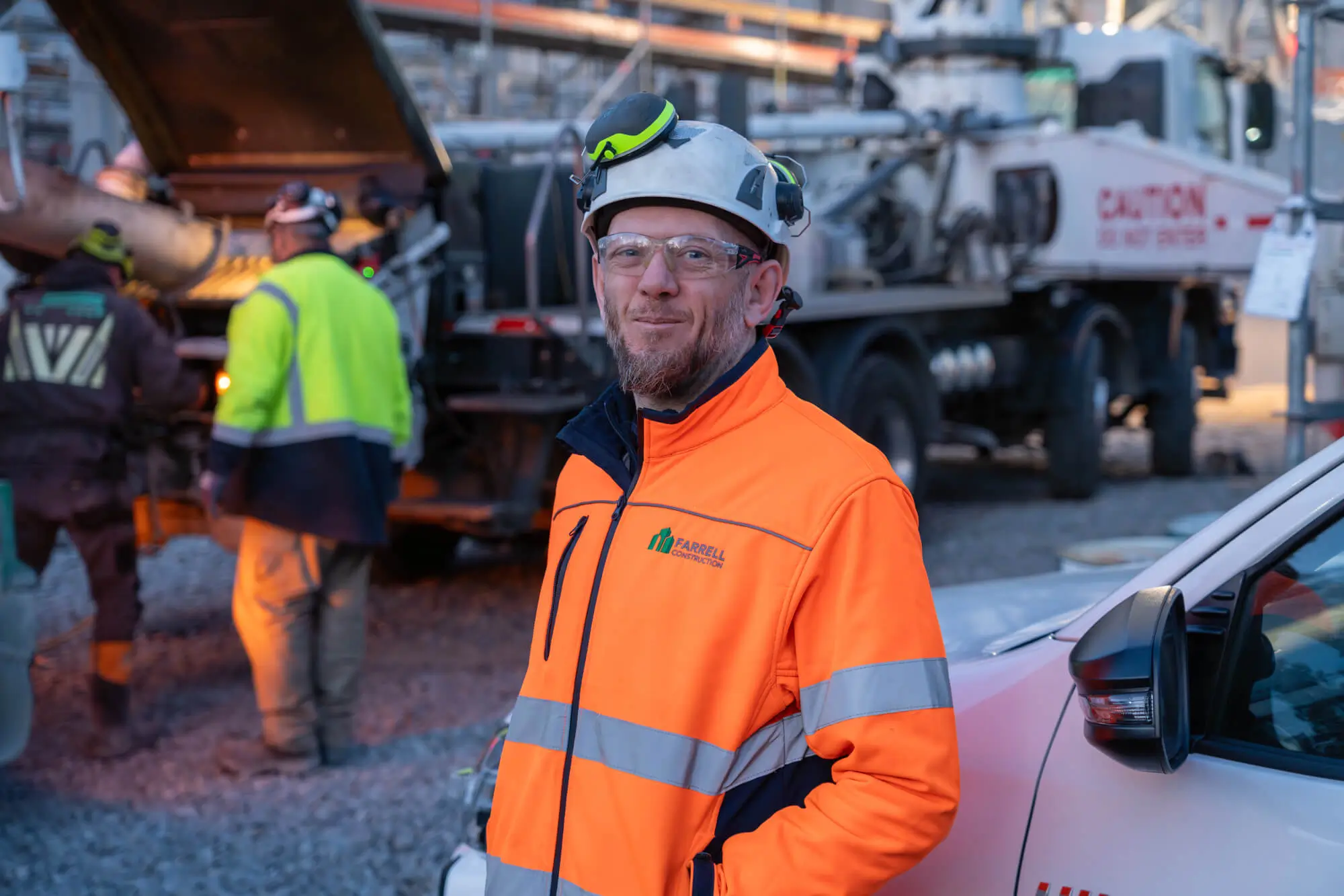 Farrell Construction worker in orange high-visibility jacket and white helmet standing near a white vehicle at a construction site.