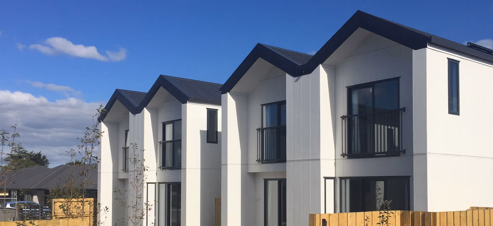 Row of newly constructed modern two-story white houses with black roofs and small balconies under a clear blue sky.