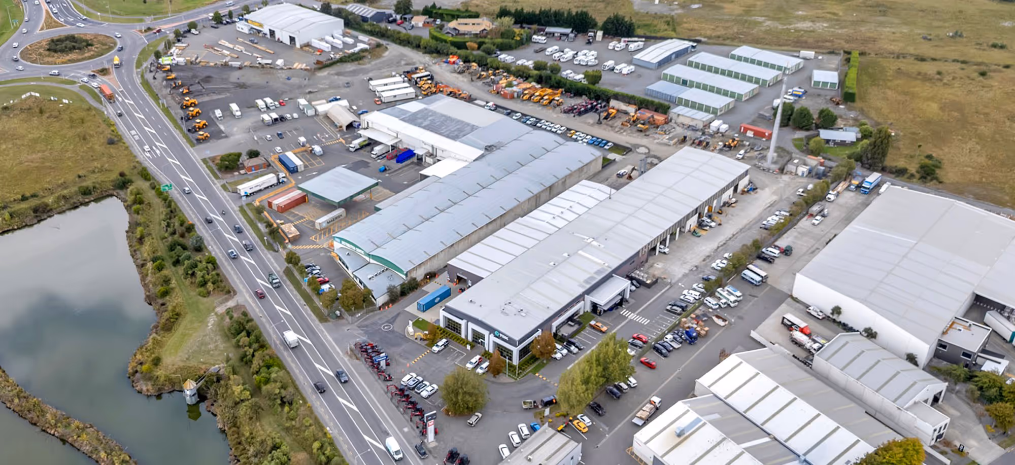 Aerial view of an industrial complex with multiple warehouses, trucks, heavy equipment, and surrounding roads near a body of water.