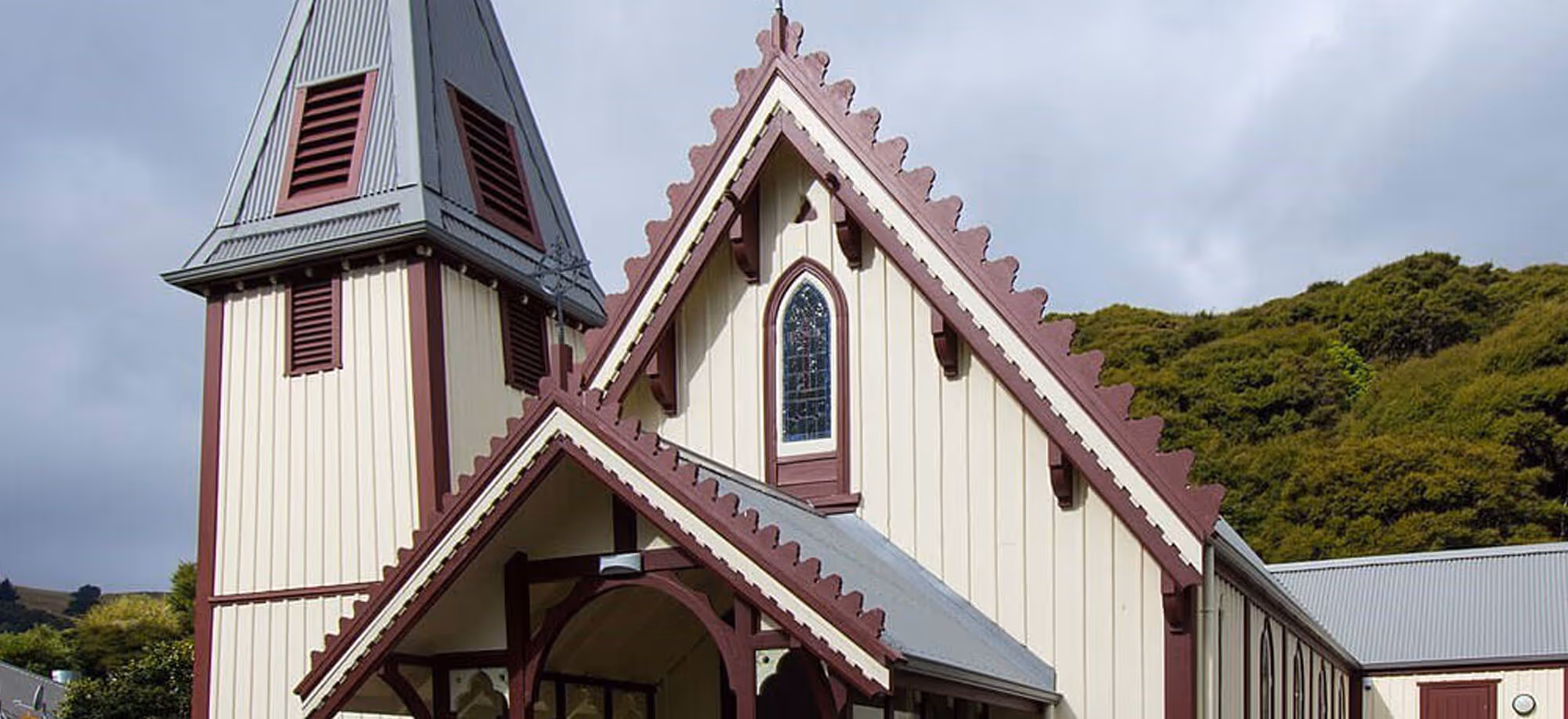 Front view of a cream and burgundy wooden church with a steep gabled roof and stained glass window in Akaroa.