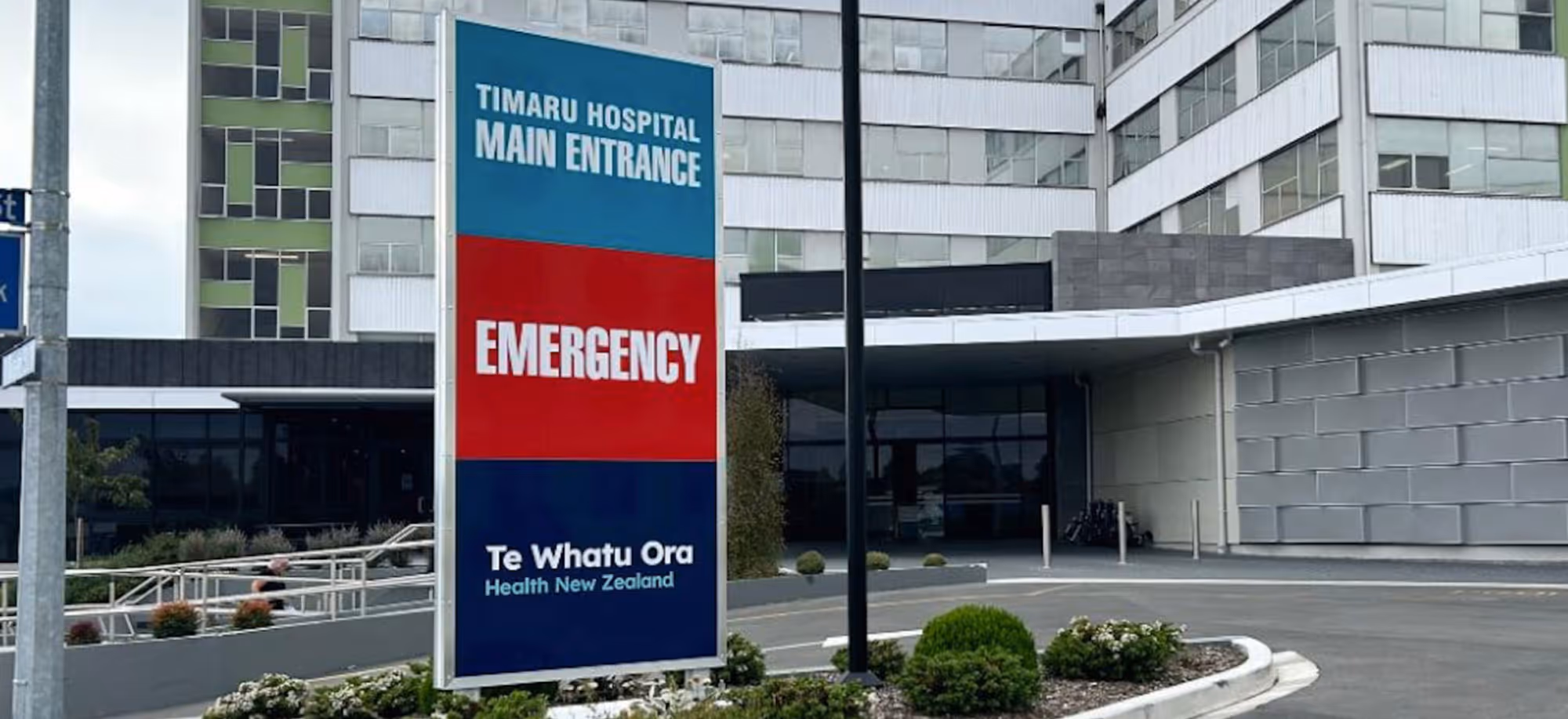 Sign at Timaru Hospital main entrance indicating emergency services and Te Whatu Ora Health New Zealand, with hospital building in the background.