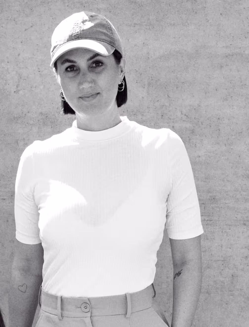 Schwarz-weiß-Fotografie einer Person mit kurz geschnittenen dunklen Haaren, die eine helle Baseballkappe, ein weißes, kurzärmliges Oberteil und eine hochgeschnittene Hose trägt; die Person steht vor einer schlichten Betonwand und blickt ruhig in die Kamera