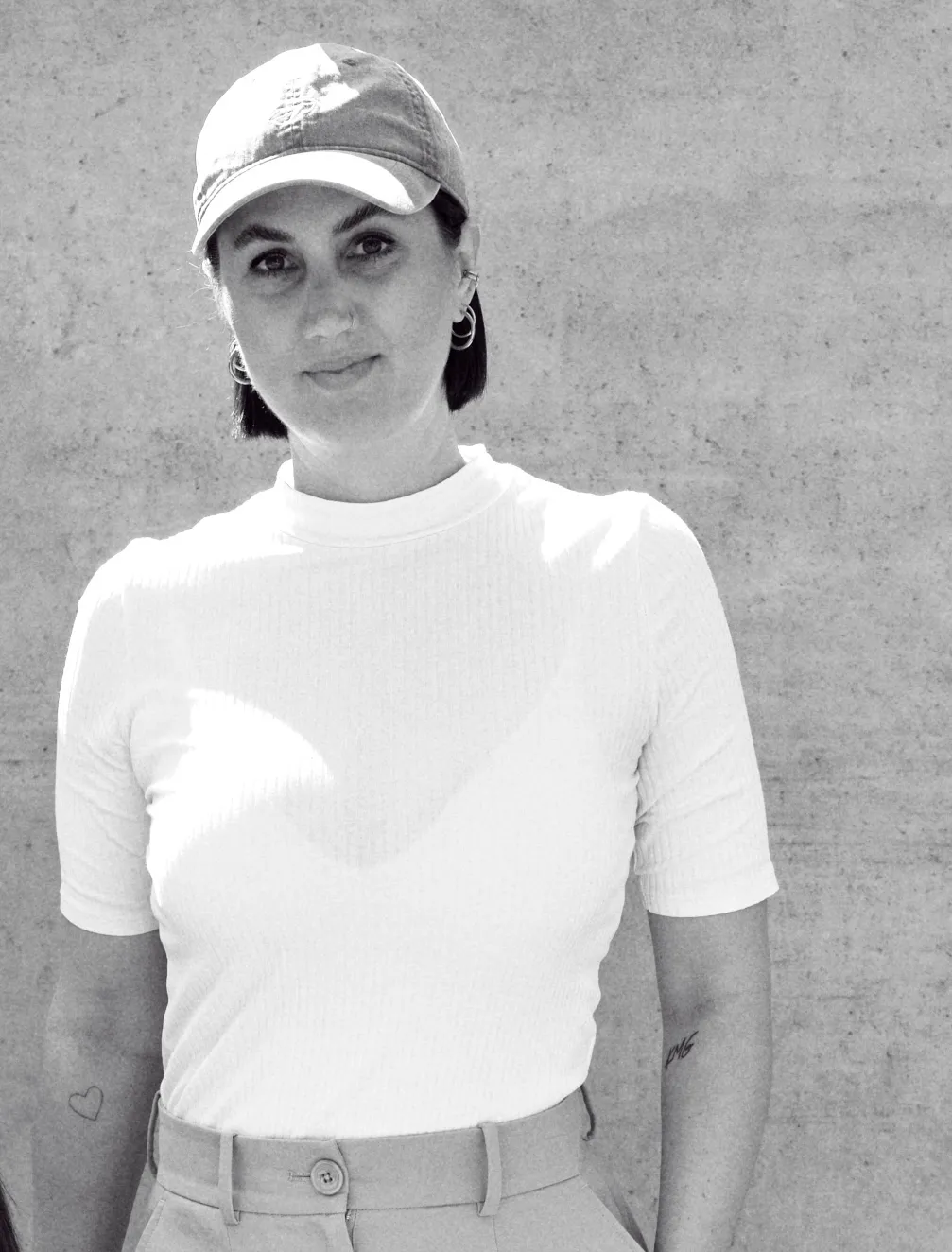 Schwarz-weiß-Fotografie einer Person mit kurz geschnittenen dunklen Haaren, die eine helle Baseballkappe, ein weißes, kurzärmliges Oberteil und eine hochgeschnittene Hose trägt; die Person steht vor einer schlichten Betonwand und blickt ruhig in die Kamera