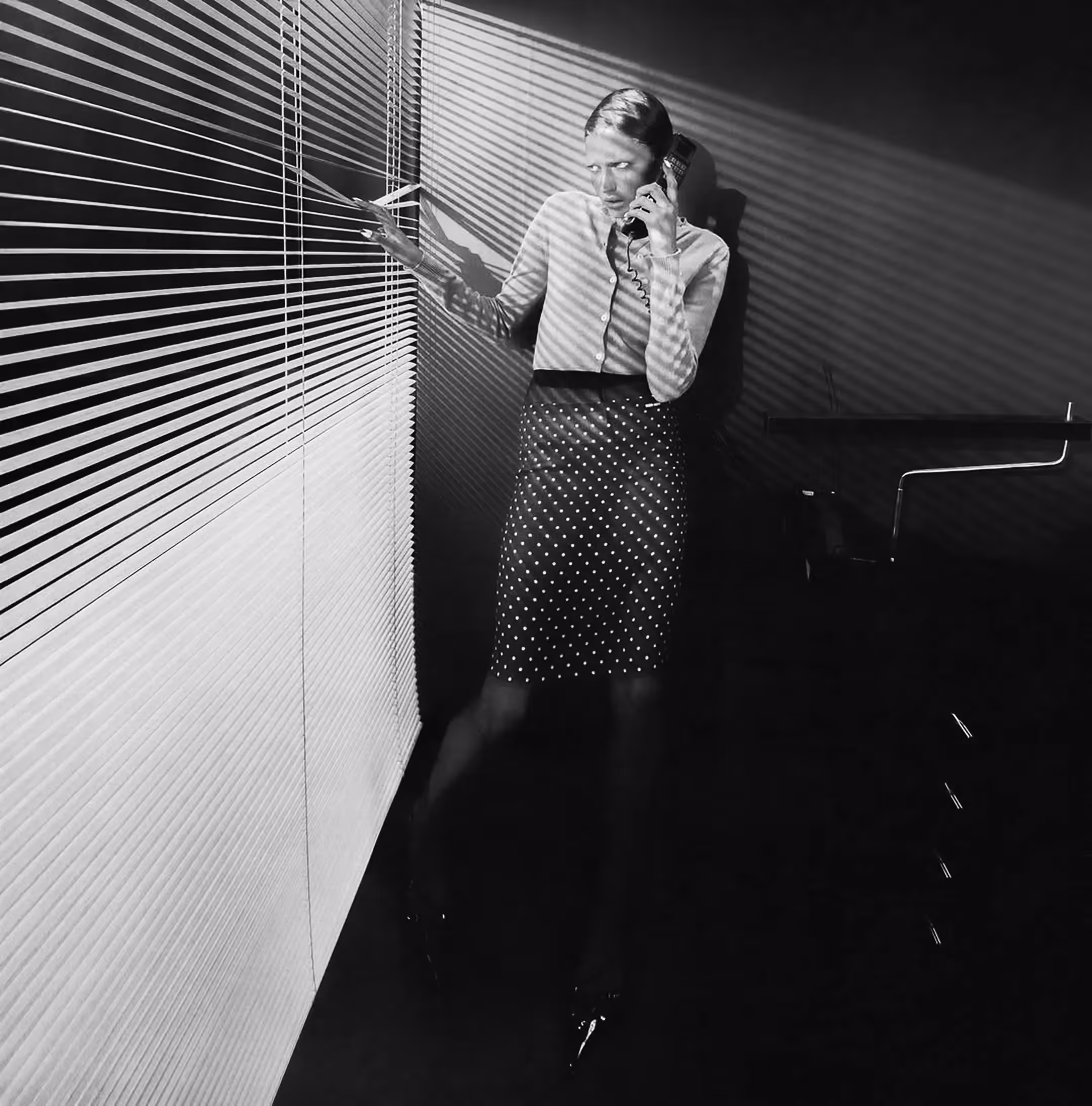 Frau im Büro telefoniert und schaut durch halb geöffnete Jalousien; Lichtstreifen zeichnen Muster auf Wand und Kleidung.