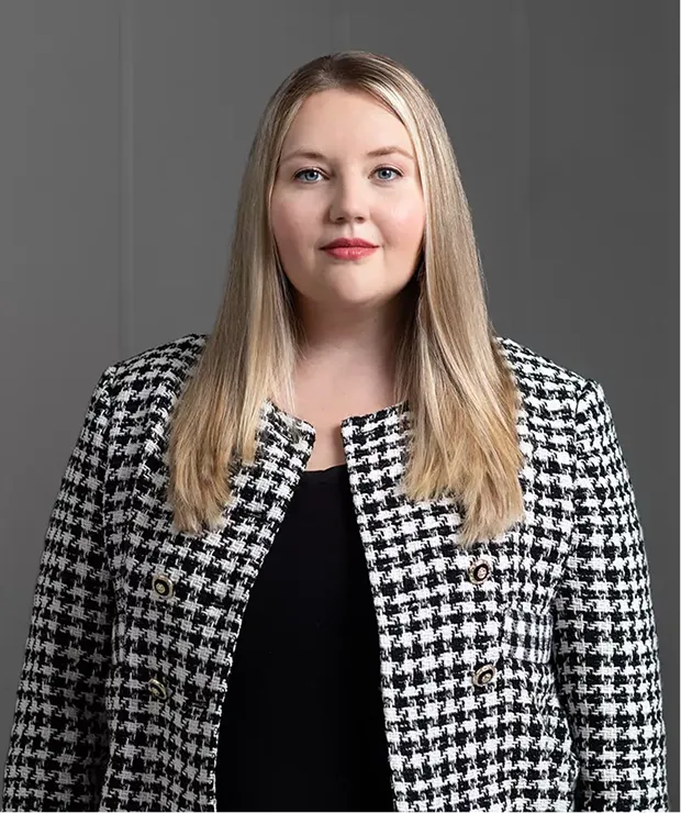 Porträtfoto einer Frau mit langen, glatten, blonden Haaren, die einen schwarz-weißen Hahnentritt-Blazer über einem schwarzen Oberteil trägt und vor einem neutral grauen Hintergrund steht.
