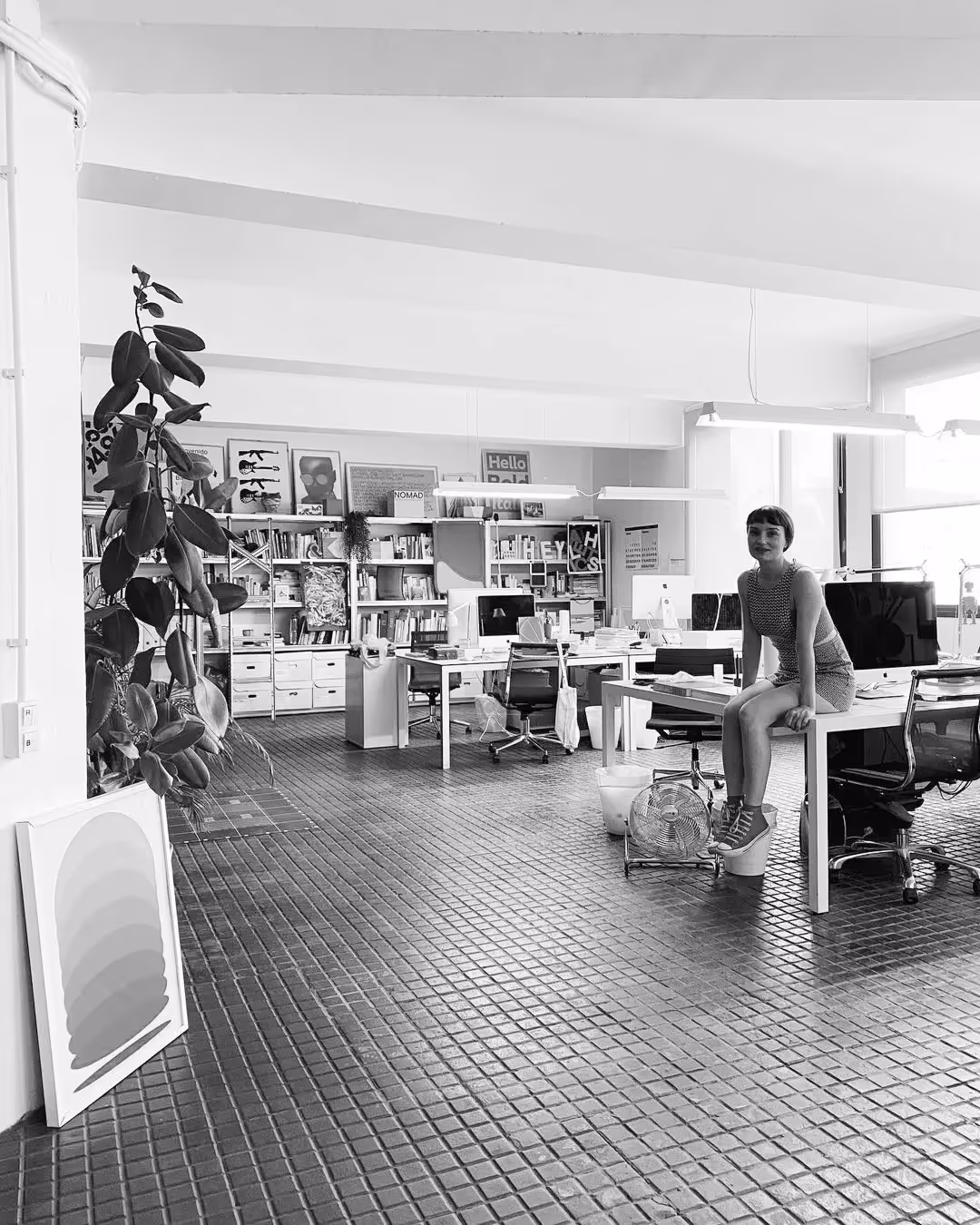 Schwarzweißaufnahme eines offenen Designbüros mit Regalwand, Pflanzen und mehreren Schreibtischen; eine Frau sitzt lächelnd auf der Tischkante nahe der Fenster.