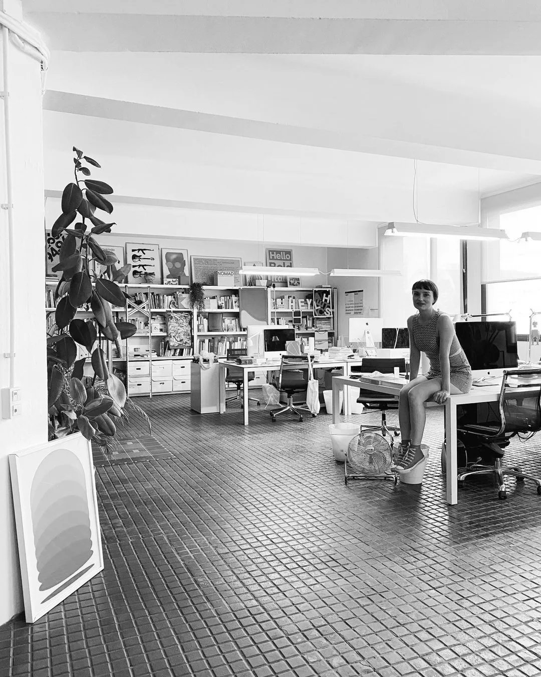 Modernes, kreatives Büro mit offener Arbeitsatmosphäre: Helle Arbeitsplätze, Bücherregale voller Inspiration und persönliche Details zeigen eine Arbeitskultur, die Design, Offenheit und Gemeinschaft lebt. Eine Mitarbeiterin sitzt entspannt auf dem Tisch – 