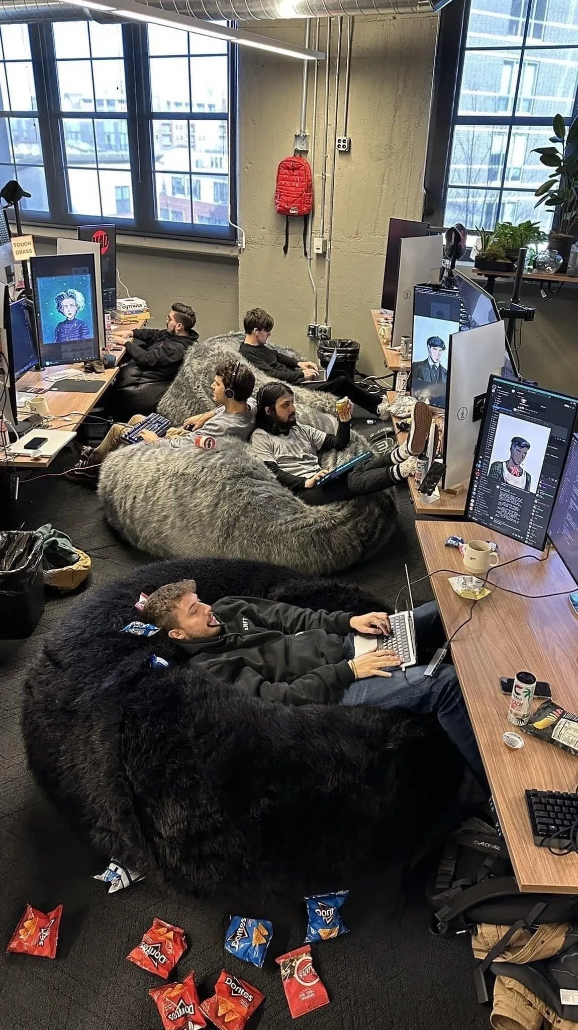 Offenes Büro mit mehreren Personen, die auf großen Sitzsäcken an PCs arbeiten; Snacks auf dem Boden, große Industrie-Fenster, lockere Startup-Atmosphäre.
