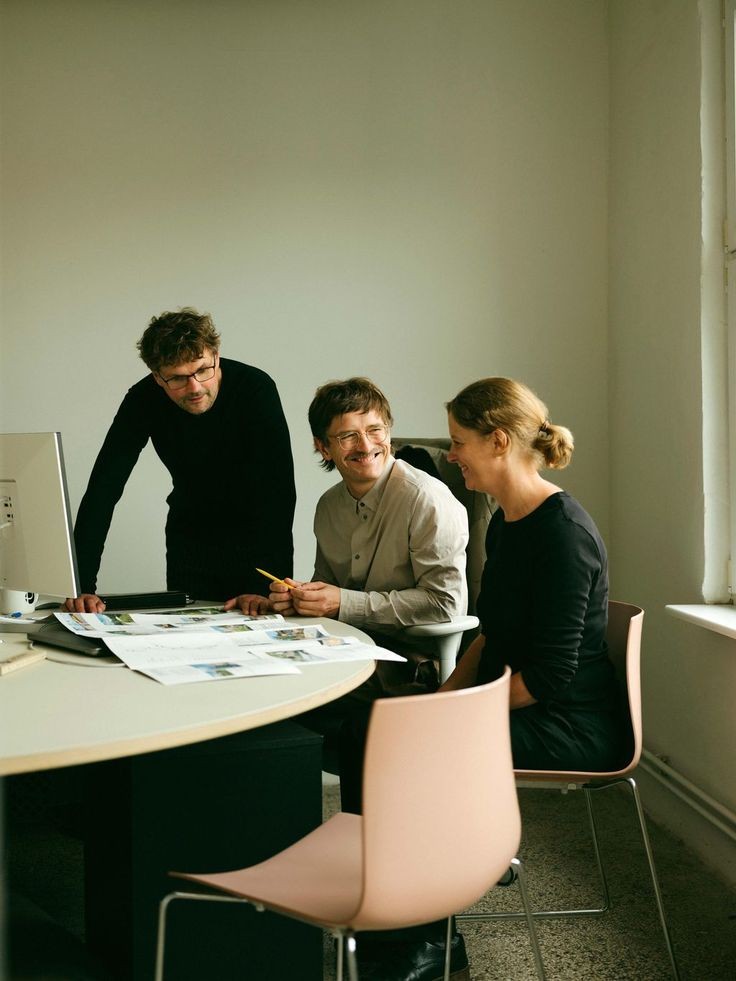 Drei Kolleg:innen an einem runden Tisch im hellen Büro, lächelnd im Gespräch über ausgedruckte Unterlagen neben einem Monitor.