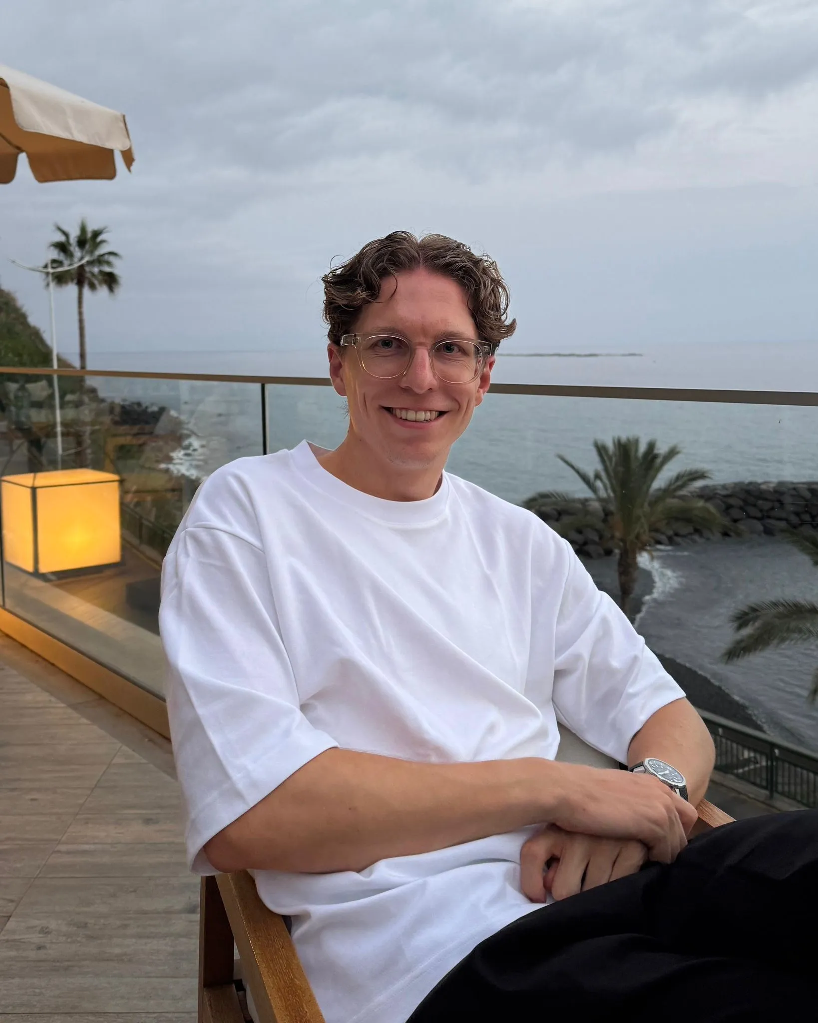 Johannes Kölmel sitzt lächelnd in einem Outdoor-Bereich mit Meerblick. Er ist Voice of stråling bei der Employer Branding Agentur stråling sowie Experte für Social Growth & Community und Influencer Marketing.