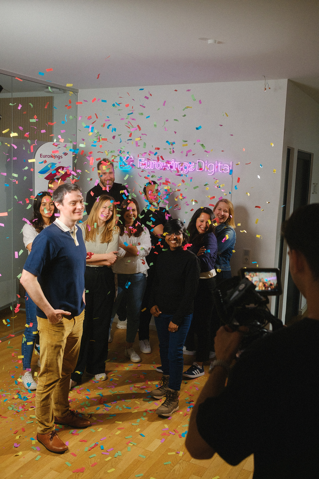 Mitarbeiterteam von Eurowings Digital feiert umgeben von buntem Konfetti, während eine Kamera den Moment festhält; lebendige Szene, wie sie von der Employer Branding Agentur stråling für authentische Team- und Kulturaufnahmen produziert wird.