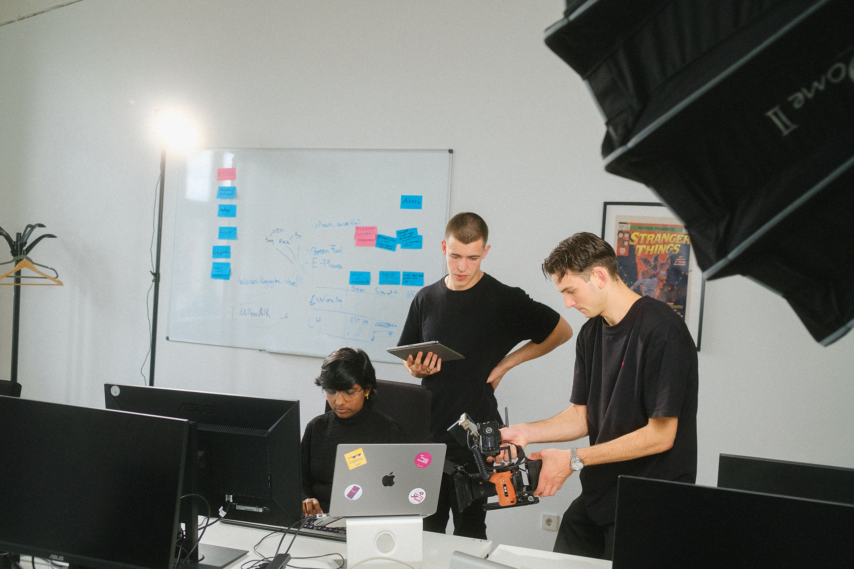 Drei Personen arbeiten gemeinsam in einem modernen Büro: Eine Person am Computer, zwei weitere mit Kamera und Tablet, während im Hintergrund ein Whiteboard mit Notizen hängt; authentische Behind-the-Scenes-Szene, wie sie die Employer Branding Agentur stråling zur Darstellung moderner Team- und Arbeitskultur produziert.