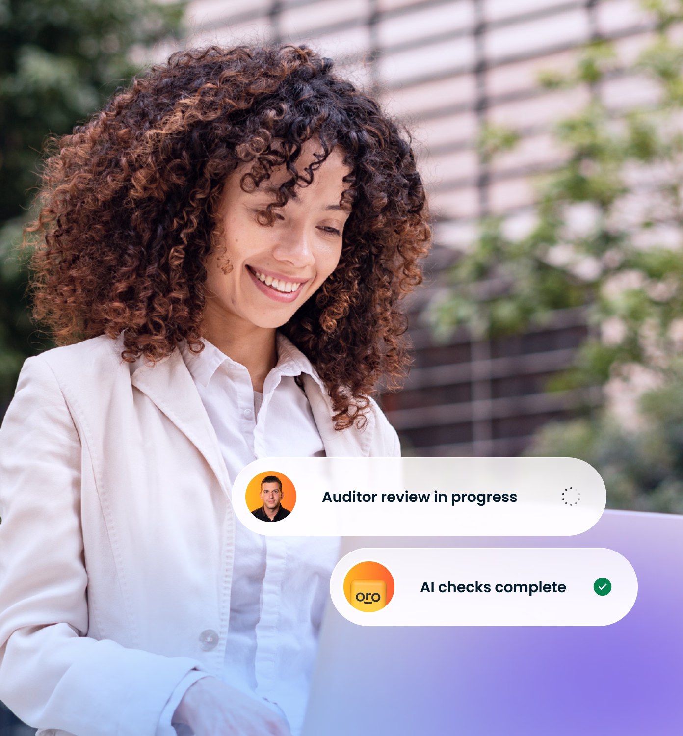 Smiling woman with curly hair wearing a white shirt and light jacket looking at a laptop, with overlay text showing a progress status reading 'Auditor review in progress' and 'AI checks complete' with checkmark.