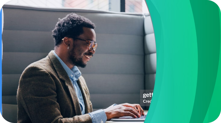 Smiling man with glasses and a blazer typing on a laptop while seated on a cushioned bench near a window.