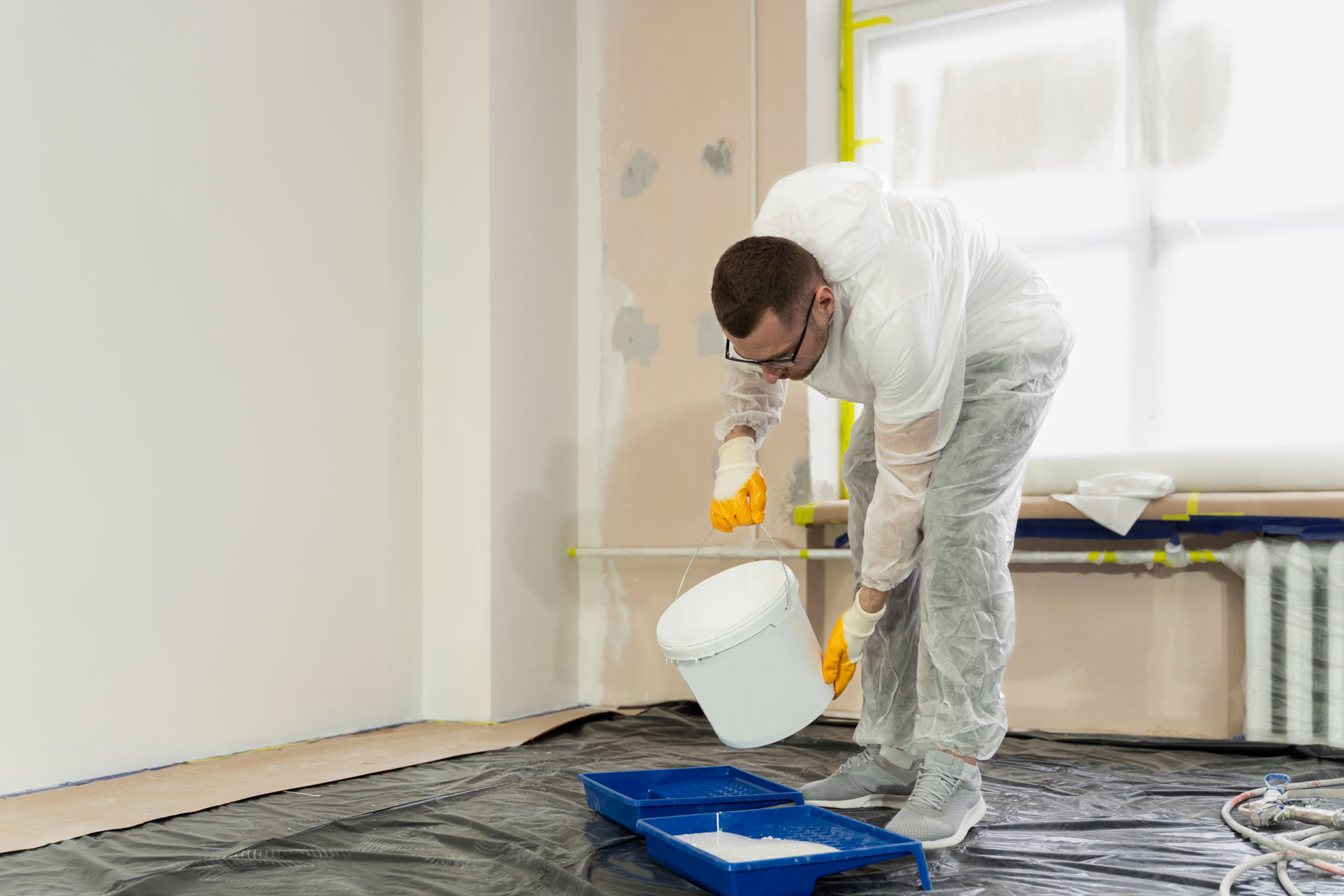 Painter pouring white paint into tray in a room.