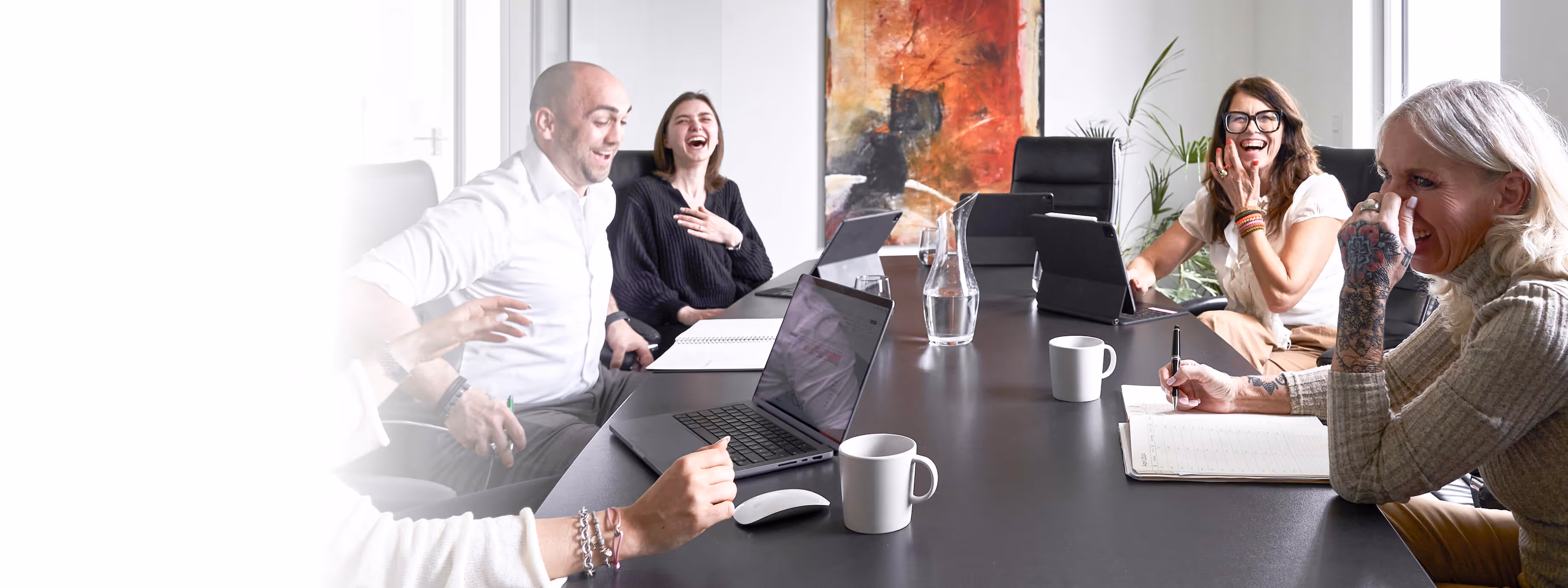 Fünf Menschen lachen und arbeiten gemeinsam an einem Konferenztisch mit Laptops und Notizbüchern.