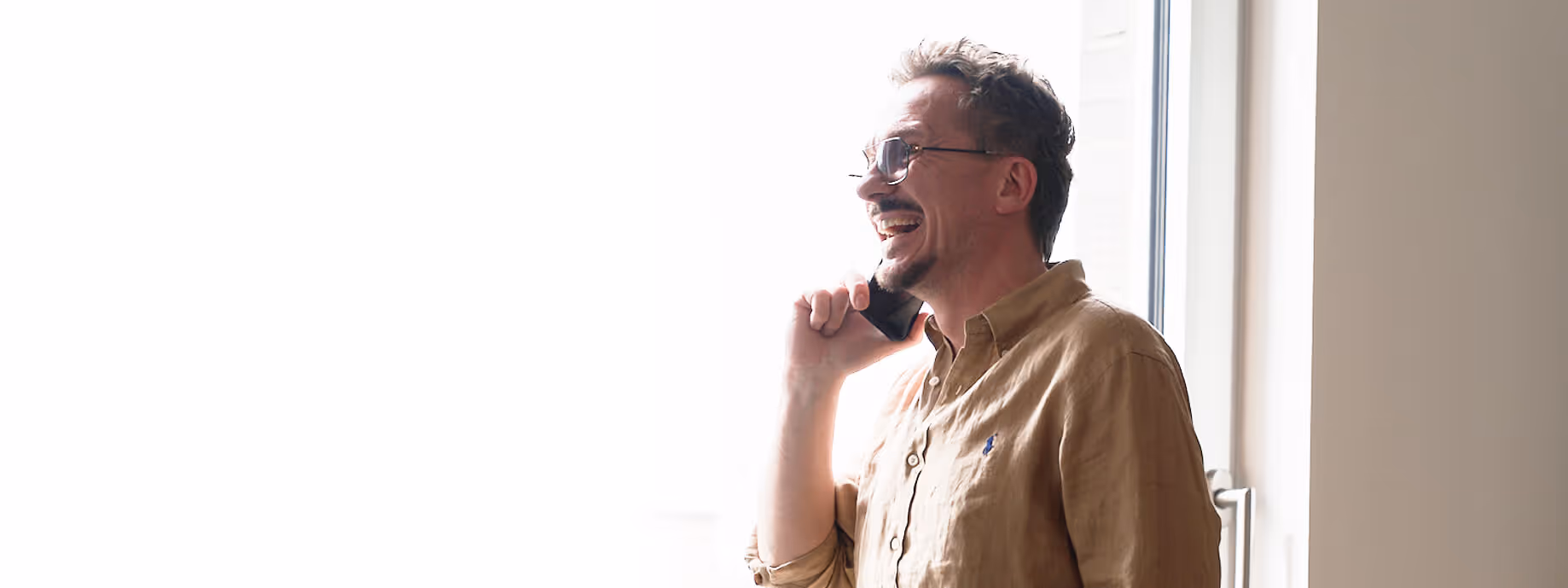Mann mit Brille und braunem Hemd telefoniert lachend am Fenster.