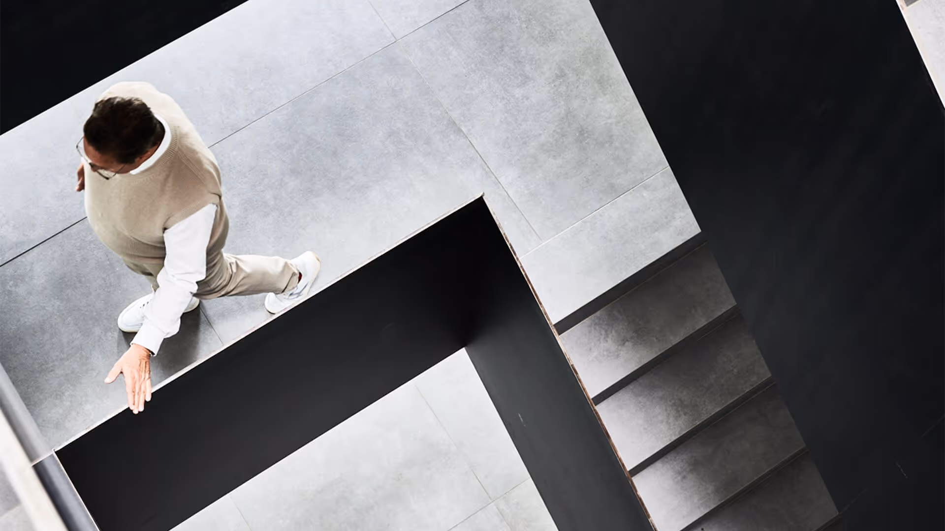 Mann mit Pullover und Brille geht eine Treppe mit grauen Stufen entlang und berührt das Geländer.
